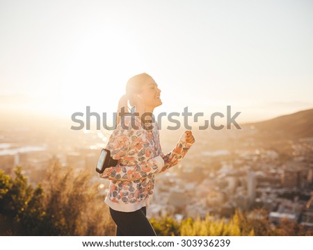 Shot of fit female runner in morning with bright sunlight. Young woman outdoors on a run smiling.