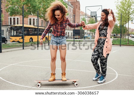 African young woman standing on a longboard with support from her friend. Woman learning to ride skateboard in a basketball court.