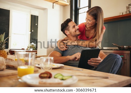 Shot of loving young couple in kitchen by breakfast table in morning. Man using digital table while woman hugging him from behind, both looking at each other smiling.