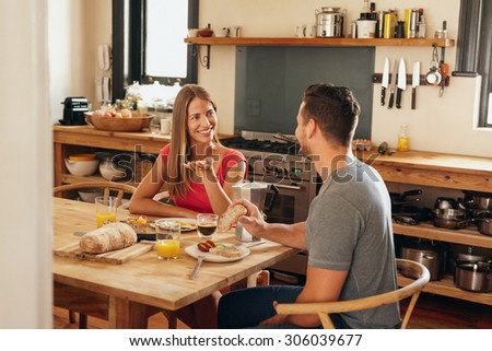 Happy young couple sitting at breakfast table in morning having a conversation. Young woman talking with her boyfriend while eating breakfast together in kitchen.