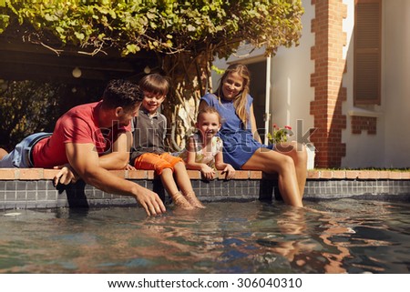 Portrait of young family with two children relaxing by their swimming pool having fun. Parents with kids sitting on the edge of pool on a sunny day.