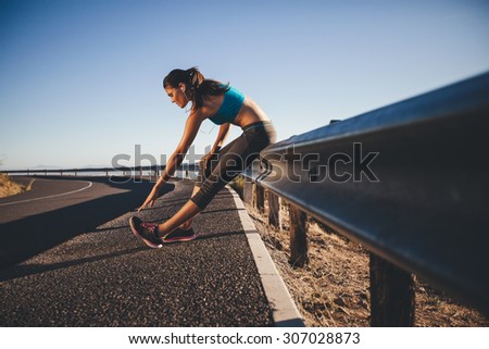 Young woman doing some stretching after a run. Runner leaning on road guardrail relaxing her calf muscles.