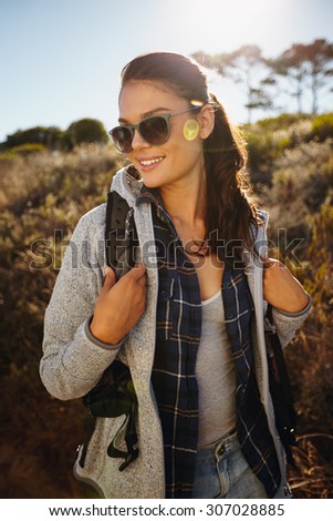 Outdoor shot of happy young woman hiking on a sunny day. Caucasian female hiker wearing sunglasses carrying a backpack looking away smiling.