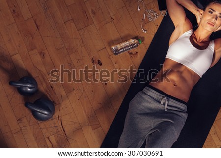 Top view of fitness model lying on exercise mat. Overhead shot of fitness instructor tired resting on mat with water bottle, mobile phone and kettle bell on floor.