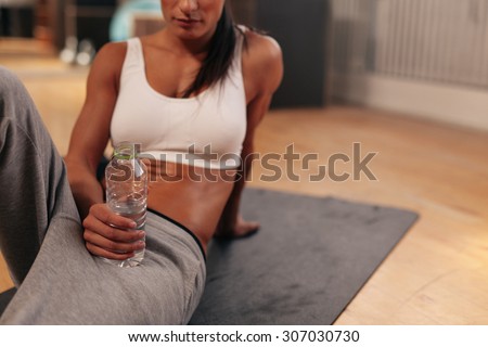 Cropped shot of relaxed young woman holding water bottle. Fitness woman in sports wear sitting on exercise mat at gym.