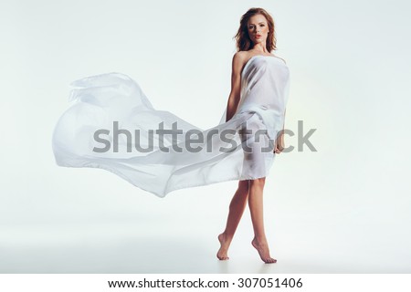Sexy nude brunette woman covered in sheer fabric on white background. Beautiful female model looking at camera.