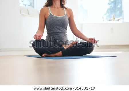 Cropped shot of fit female sitting cross legged on exercise mat with hands on knees meditating. Woman in lotus pose at health club. Padmasana, meditation posture.