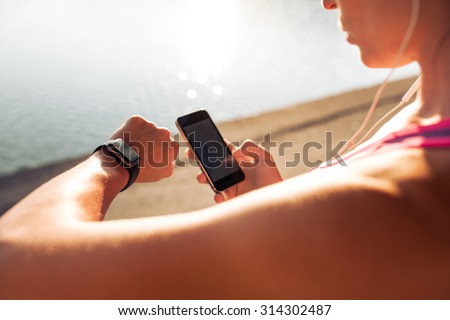 Sportswoman looking at smartwatch and holding smart phone in her other hand, outdoors. Fitness female setting up her smartwatch for her run.