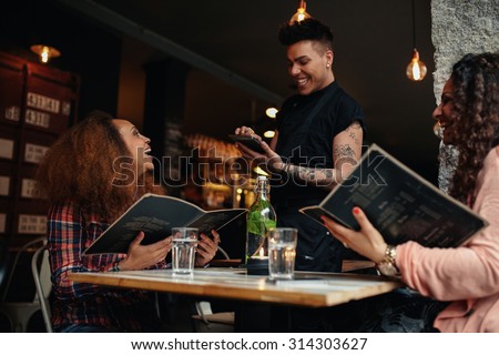 Cheerful two young women sitting at cafe holding menu card giving order to waiter. Young woman placing order to a waiter at restaurant while sitting with her friend.