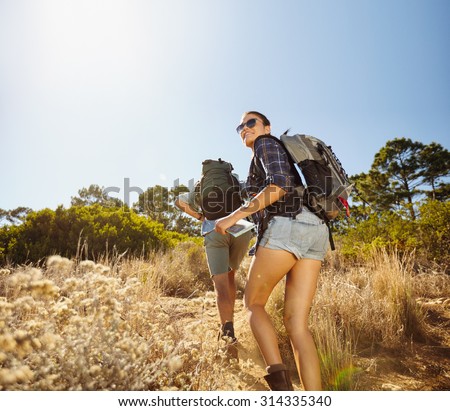 Young people doing a uphill climb while hiking in countryside. Woman with map looking back over her shoulder smiling with man walking in front. Couple on hiking trip.
