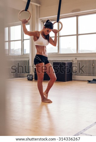 Full length shot of fit young woman resting after exercising on gymnastic rings. Female in sports wear standing in gym holding rings and looking down.