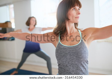 Fitness trainer doing the warrior pose during yoga class. Yoga teacher performing Virabhadrasana position in gym with people n background.