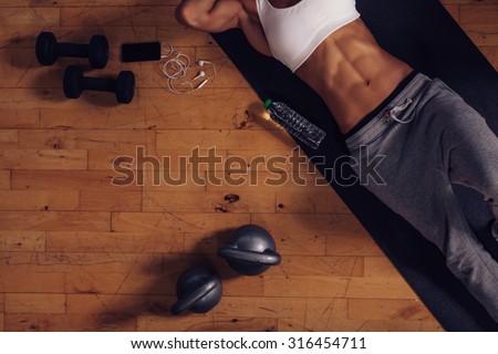 Top view of young woman lying on yoga mat in gym. Fitness woman relaxing after exercise session with a water bottle, dumbbells and kettlebell on floor.