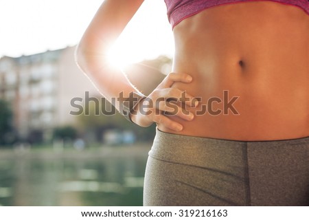 Mid section of fit woman\'s torso with her hands on hips. Female runner wearing smartwatch device with sun flare, outdoors.