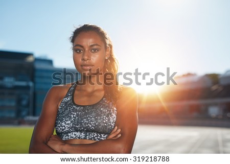 Portrait of confident young sportswoman standing with her hands folded on athletics stadium looking at camera with bright sunlight from behind. African female athlete on race track.