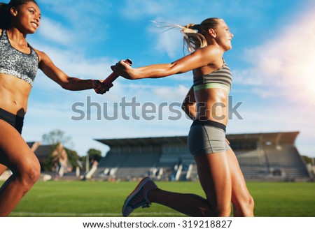 Female athletes passing over the baton while running on the track. Young women run relay race, track and field event.