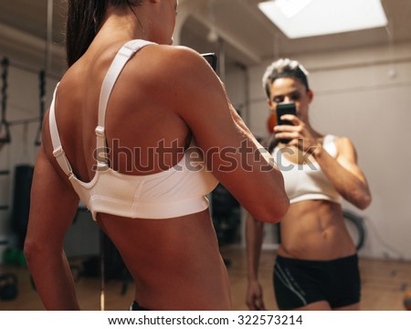 Sporty young woman taking a picture of herself in a mirror. Fitness model taking a selfie in front of a mirror in gym.