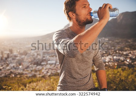 Young runner taking a break after morning run drinking water. Young man relaxing after a running training session outdoors in countryside on sunny day.