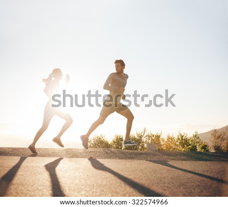 Young people running on country road with bright sunlight. Outdoor shot of young man and woman jogging in morning.