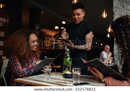 Happy young people sitting in restaurant reading menu card and giving orders to the male waiter, while waiter putting order onto a digital tablet.