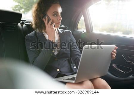 Businesswoman sitting on back seat of car with laptop talking on mobile phone. Female executive travelling to work in luxury car.