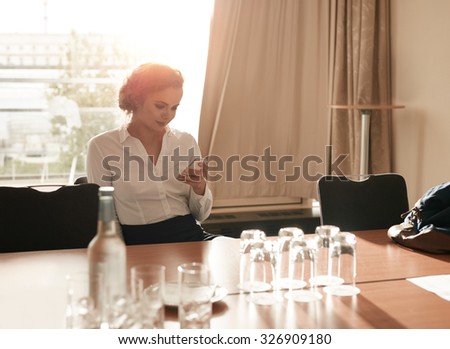Portrait of young businesswoman sitting in business conference room using mobile phone. Female executive sitting in hotel conference room reading text message on her mobile phone.
