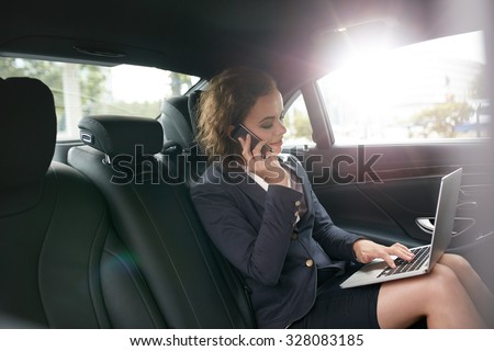 Businesswoman with laptop receiving a phone call on the backseat of a car. Female entrepreneur working during travelling to office in a luxury car.