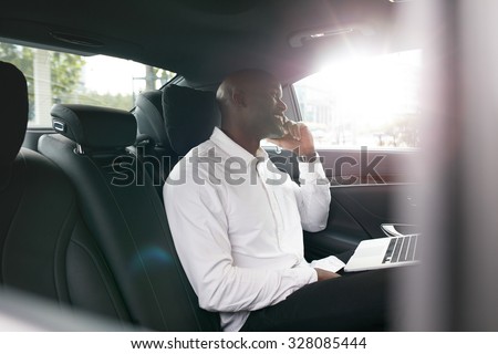 African businessman with laptop talking on mobile phone inside a car. Young entrepreneur working during travelling to office in a luxury car.
