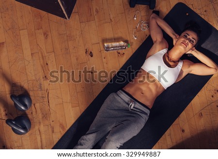 Top view of relaxing young woman lying on fitness mat. Overhead shot of female athlete resting after intense workout with water bottle, mobile phone and kettle bell on floor.