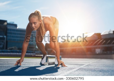 Professional female track athlete in set position on sprinting blocks of an athletics running track. Runner is in a athletics stadium with bright sunlight.