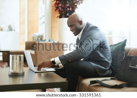 Happy businessman using laptop in hotel lobby. African business executive sitting on couch at hotel lobby working on his laptop and smiling.