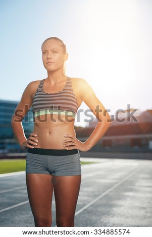 Female runner standing with her hands on hips looking away. Determined woman athlete on race track in athletics stadium with sun flare.
