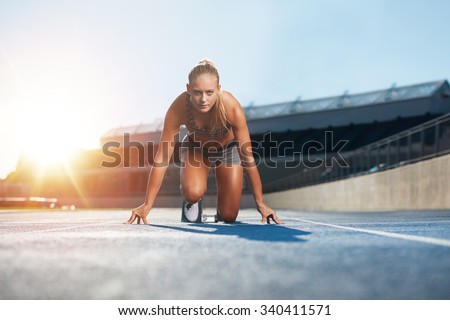 Confident young female athlete in starting position ready to start a sprint. Woman sprinter ready for a run on racetrack with sun flare.