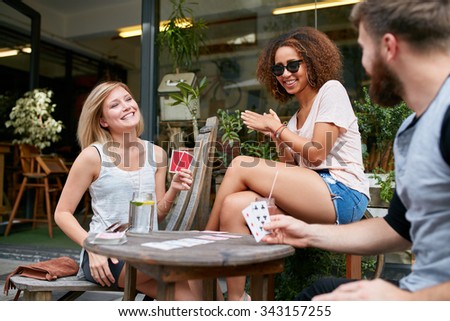 Three friends sitting in outdoor cafe and playing cards and having fun. Happy young people at sidewalk coffee shop enjoying playing poker game.