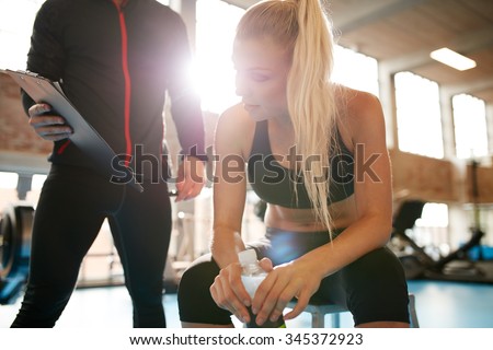 Young woman taking a break from her workout and looking at her fitness plan on clipboard. Female with her personal trainer at gym.