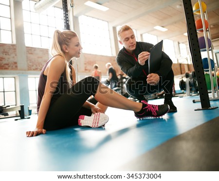 Personal trainer helping young woman on her work out routines in gym. Female sitting on floor with her personal trainer showing fitness report on a clipboard.