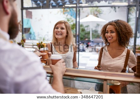 Indoor shot of two young women friends smiles as they receive their hot drinks from the cafe counter. Happy young female friends buying coffee.