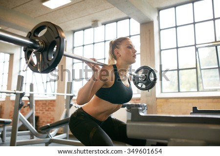 Female exercising in gym doing squats with extra weight on her shoulders. Young woman working out with heavy weights in a fitness club.