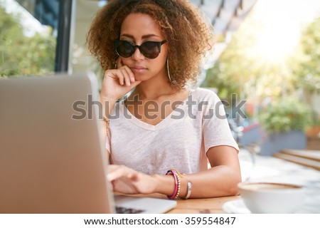 Outdoor shot of young woman at sidewalk cafe reading emails on her laptop. Stylish young girl sitting at outdoor coffee shop using laptop.