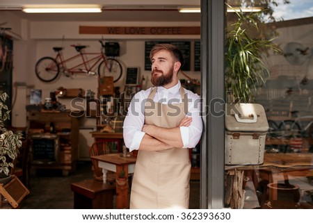 Portrait of a cafe owner standing in the doorway of his coffee shop. Young waiter standing with his arms crossed at the door of a restaurant and looking away.