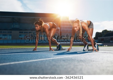 Two female athletes at starting position ready to start a race. Sprinters ready for race on racetrack with sun flare.