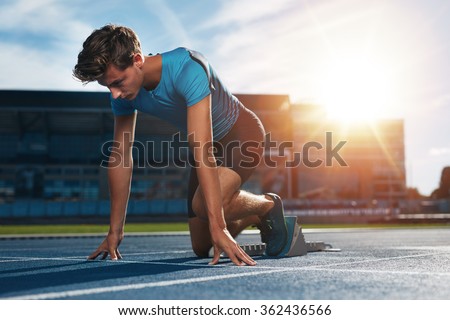 Young male athlete at starting block on running track. Young man in starting position for running on sports track. Sprinter about to start a race at stadium with sun flare.