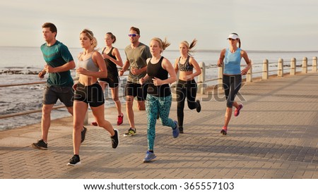 Portrait of young people running along the beach boardwalk by the ocean. Fit young men and women running training outdoors by the seaside.