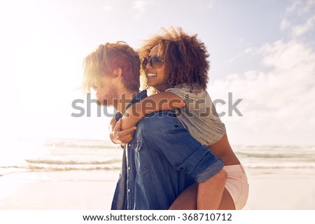 Side view portrait of young man carrying his girlfriend on his back at the beach. Boyfriends giving piggyback ride to his beautiful girlfriend at seashore.