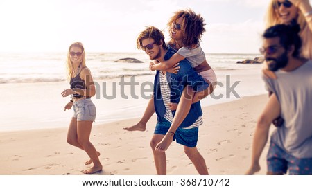 Two happy young men giving their girlfriends piggyback rides at the beach. Group of friends enjoying beach holidays.