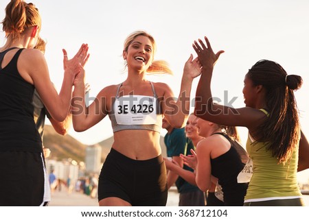 Multi ethnic group of young adults cheering and high fiving a female athlete crossing finish line. Sportswoman giving high five to her team after finishing the race.