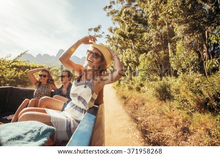 Portrait of a cheerful young woman sitting in the back of pickup truck with friends. Young people enjoying on a road trip.