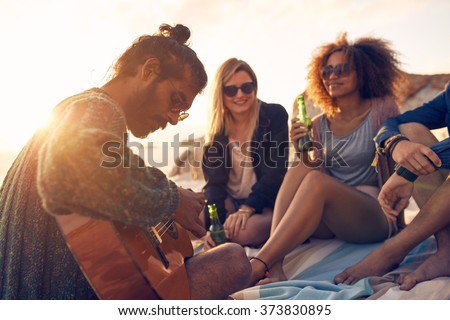 Hipster playing guitar for friends at the beach. Group of young people drinking beer and listening to music.