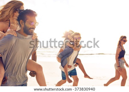 Group of friends having fun on the beach, Young men piggybacking women on the sea shore. Mixed race young people enjoying summer vacation.