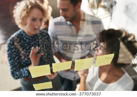 Three business people having a meeting in office. They are standing in front of glass wall with post it notes and discussing.